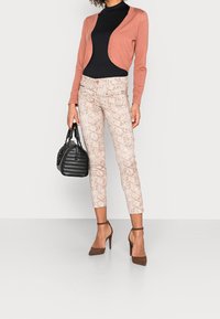 Kvinna i sicksacksmönstrade cropped byxor, svart topp, rosa cardigan, bruna klackar och bär en svart handväska framför en neutral bakgrund.