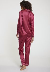 Zijdeachtige rode pyjama set met een lange mouwen button-up top met kraag en bijpassende broek. Witte piping accenten bij de manchetten en kraag.
