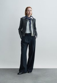 Γκρι σακάκι με κουμπιά σε αποχρώσεις, πάνω από ένα ανοιχτό γκρι πουλόβερ, σε συνδυασμό με ψηλόμεσα παντελόνια με λεπτές ρίγες και ένα φουλάρι με σχέδια. Μαύρα παπούτσια.