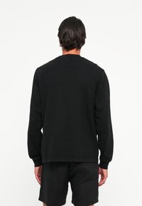 Man met donker haar in een eenvoudig zwart shirt met lange mouwen en zwarte shorts, staand met de rug naar de camera tegen een witte achtergrond.