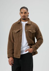 Brown fleece jacket with a textured fabric, button-up front, two chest pockets, and a collar, layered over a white shirt.