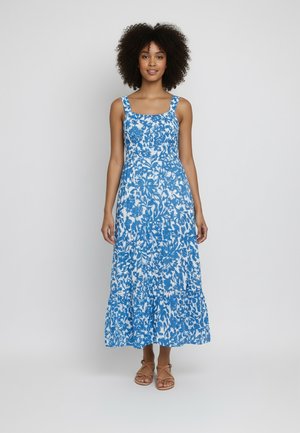 Donna con capelli ricci che indossa un vestito maxi senza maniche a fiori blu e bianchi e sandali marroni, in piedi davanti a uno sfondo neutro.