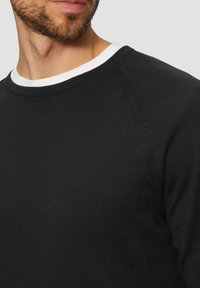 Zwarte trui met een witte ronde hals. De stof lijkt soepel met ribbelstructuur langs de schouders en de halslijn.
