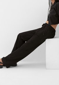 Pantalon noir à coupe décontractée avec taille élastique, poches latérales et tissu texturé. Porté avec une chemise noire cropped à boutons.