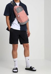 Zaino rosa e grigio con una forma arrotondata, texture in tessuto, tasca anteriore con zip e tracolla regolabile. Indossato da una persona in abbigliamento casual.