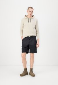 Felpa beige con cordini, pantaloni cargo neri con tasche e stivali marroni con lacci. Modello in piedi contro uno sfondo bianco.