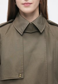 Olivgrön trenchcoat med hög krage, med knapp- och sömdetaljer, gjord av ett slätt, strukturerat tyg.