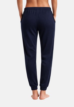 Personne au dos nu portant un pantalon de jogging bleu marine avec des chevilles retroussées, debout pieds nus avec les mains dans les poches, tournée vers l'arrière.