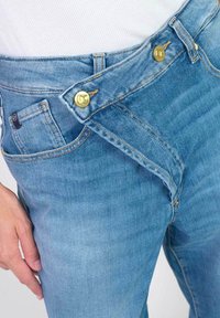 Des jeans en denim bleu clair présentent un design crossover unique avec des boutons en métal doré, des coutures visibles et des poches avant.