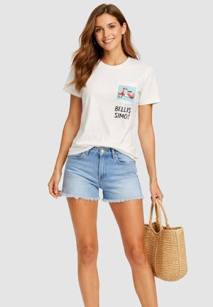 Donna sorridente con lunghi capelli castani che indossa una maglietta bianca con grafica di uno scooter e testo, pantaloncini di jeans, e tiene una borsa tote intrecciata di paglia.