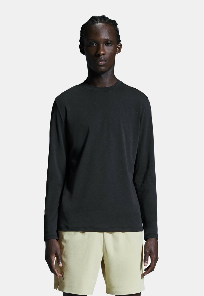 Schwarzes Langarmshirt aus glattem Stoff, kombiniert mit hellgelben Shorts. Das Shirt hat einen runden Ausschnitt und eine lässige Passform.