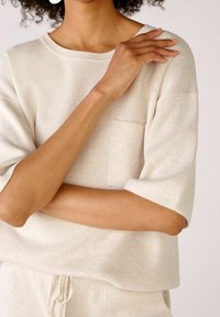 Žena v béžovém úpletu s krátkým rukávem a kapsičkou na prsou, s překříženýma rukama a dotýkající se ramene, zobrazená pouze od pasu nahoru a krk.