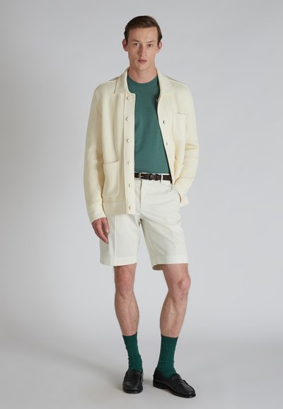Cardigan a costine beige chiaro con bottoni, t-shirt verde, pantaloni corti bianchi, calzini verdi e mocassini neri; design minimalista e vestibilità classica.