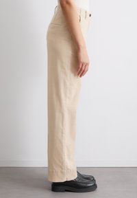 Pantalons beige taille haute à jambe droite, en coton, texture subtile, fermeture à un bouton, poches latérales, ourlet retroussé, portés avec des chaussures noires.