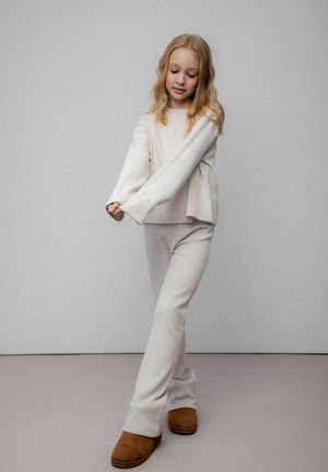 Young girl with blonde hair wears white ribbed knit sweater and pants, standing with crossed legs and brown slippers on gray background.