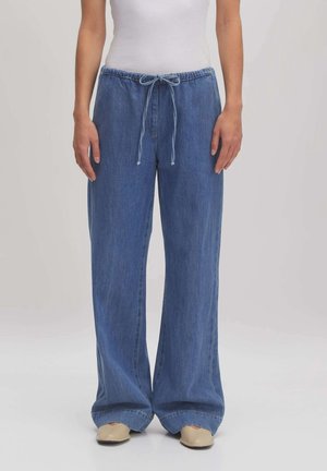 Frau trägt lockere blaue Jeanshosen mit Kordelzug an der Taille und beige Schuhe, steht vor einem schlichten Hintergrund.