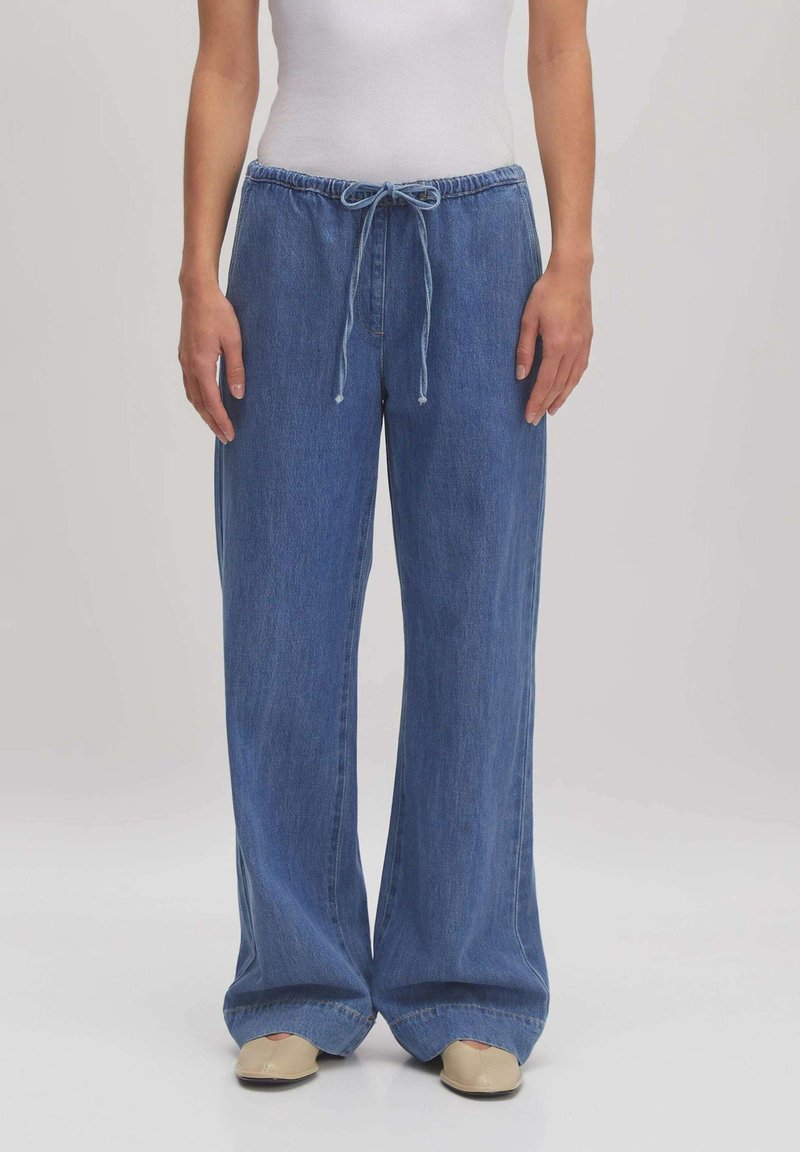 Femme portant un pantalon en jean bleu large avec une taille à cordon et des chaussures beiges, debout devant un fond uni.
