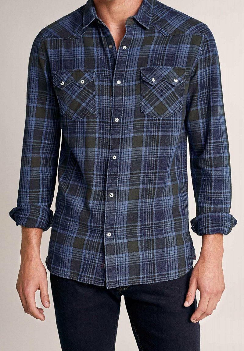 Homme portant une chemise à carreaux bleus et noirs à manches longues, avec des poches poitrine boutonnées et des manches retroussées.