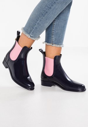 Ankle-high glossy black boots with pink elastic side panels worn with cropped frayed-hem blue jeans on a white floor.