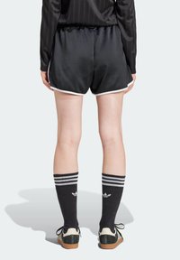 Person i svarta sportshorts med vita detaljer, svarta Adidas-strumpor med vita ränder och svarta Adidas-sneakers, står med ryggen mot betraktaren.