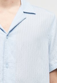 Lyseblå kortermet skjorte med vertikale striper, myk tekstur, spredd krage og to knapper foran.