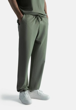 Mies pukeutunut oliivinvihreisiin kiristysnahkihin ja valkoisiin lenkkareihin, kädet taskuissa, seisoo valkoista taustaa vasten.