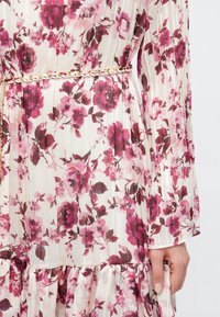 Το χέρι μιας γυναίκας ακουμπάει στο πλάι, φορώντας ένα λευκό φόρεμα με ροζ και βουργουνδί φλοράλ σχέδιο και μια λεπτή λευκή και χρυσή αλυσίδα ζώνη στη μέση.