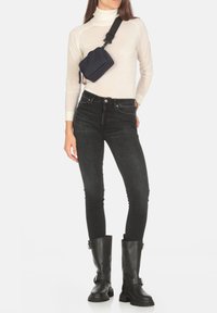 Jeans skinny neri, dolcevita crema e stivali neri fino al ginocchio. Una piccola borsa a tracolla blu navy è indossata sulla spalla. Texture lisce.