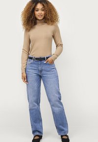Beige coltrui met lange mouwen, gecombineerd met lichtblauwe high-waisted broek met wijde pijpen en een zwarte riem, gedragen met zwarte schoenen.