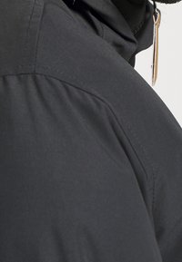 Primo piano di una persona che indossa una giacca nera con cuciture visibili e un tirazip beige che pende vicino al colletto.
