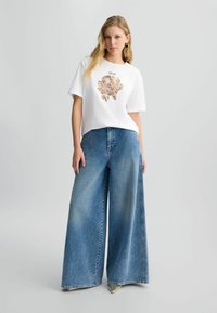 Maglietta bianca a maniche corte con design a stampa leopardata, abbinata a jeans blu a gamba larga e scarpe con dettagli metallici.