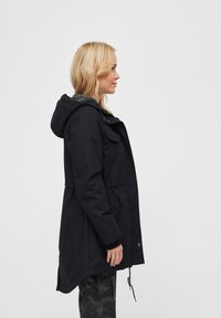 Czarna parka z kapturem, regulowanym pasem na sznurek, przednimi kieszeniami oraz miękką, szarą podszewką. Materiał wydaje się trwały o gładkiej fakturze.