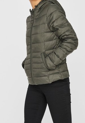 Osoba ubrana w oliwkowo-zieloną pikowaną kurtkę puchową z kapturem i bocznymi kieszeniami, w połączeniu z dopasowanymi czarnymi spodniami.