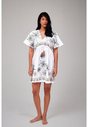 Vrouw staat blootsvoets en draagt een wit kort jurkje met groene palmboom patronen en een geknoopte kwast in de taille.