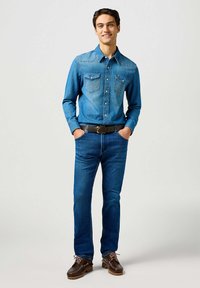 Lichtblauwe denimshirt met twee borstzakken en versterkte stiksels. Gecombineerd met blauwe jeans en bruine schoenen met veters.