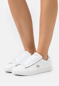 Baskets blanches en cuir avec une finition texturée, présentant un accent argenté sur le côté et les lacets, ainsi qu'un logo sur la languette.