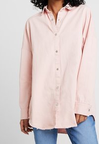 Ljusrosa långärmad skjorta med knappar och krage, med silverfärgade knappar och en något trasig fåll, tillverkad av mjuk bomull.