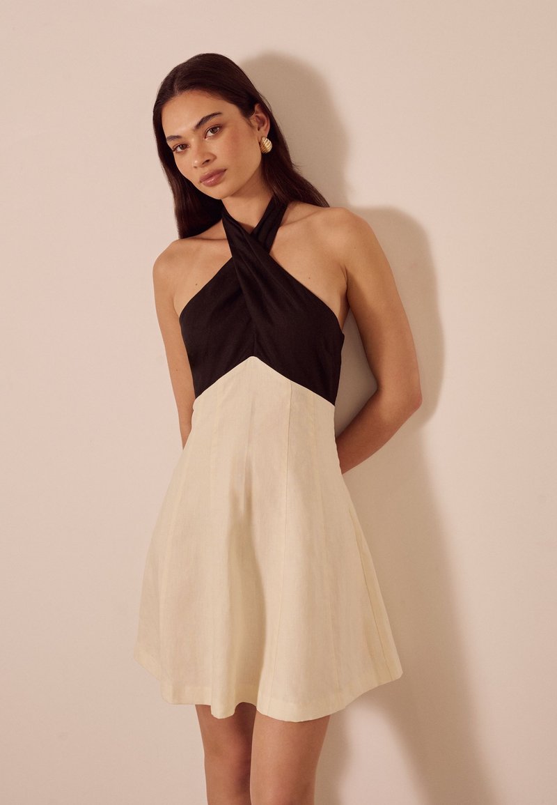 Junge Frau in einem ärmellosen Halterkleid mit schwarzem Oberteil und beiger Rock, steht vor einer schlichten hellen Wand und trägt goldene Ohrringe.