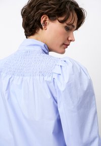 Light blue blouse with smocked back detail and puffed sleeves. Smooth cotton material, structured collar. No visible patterns or accents.