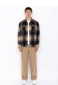 Rutet flanellskjorte med svarte, gule og grå farger, med to brystlommer og knapper. Brukt over en hvit t-skjorte og kombinert med beige bukser.