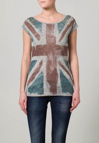 Grå bomullströja med en blek Union Jack-tryck, korta ärmar och dekorativa gyllene nitar på axlarna. Bärs med blå jeans.