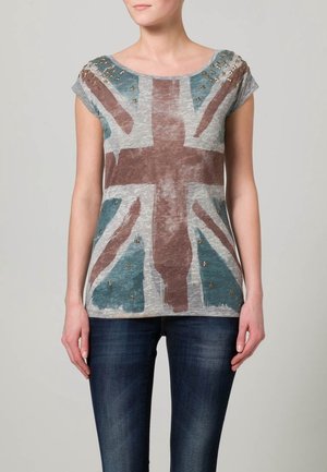 Grijze katoenen t-shirt met een vervaagde Union Jack-print, korte mouwen en decoratieve gouden studs op de schouders. Gekleed met blauwe jeans.