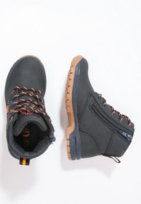 Stivaletti neri con lacci arancioni in evidenza, zip laterale e suole in gomma beige, mostrati in vista superiore e laterale su uno sfondo bianco.