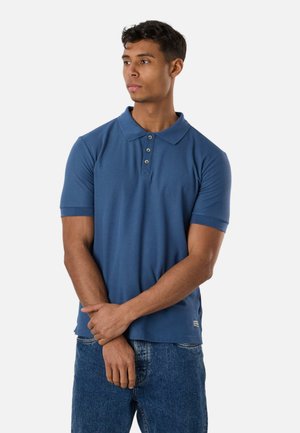 Ung mand i en blå kortærmet poloshirt og jeans, der står med armene krydsede og kigger sidelæns mod en ensartet hvid baggrund.
