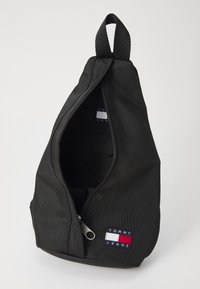 Sort stof sling tas med en lynlås åbning, der har et lille Tommy Jeans logo patch i rød, hvid og blå nær lynlåsen.
