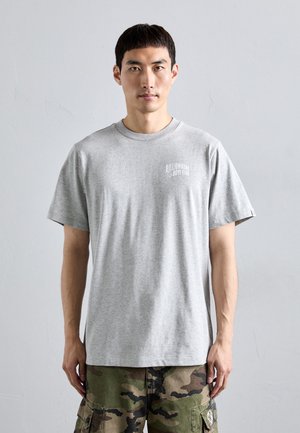 Man wearing a light gray Billionaire Boys Club t-shirt and camouflage cargo pants standing against a plain light gray background.