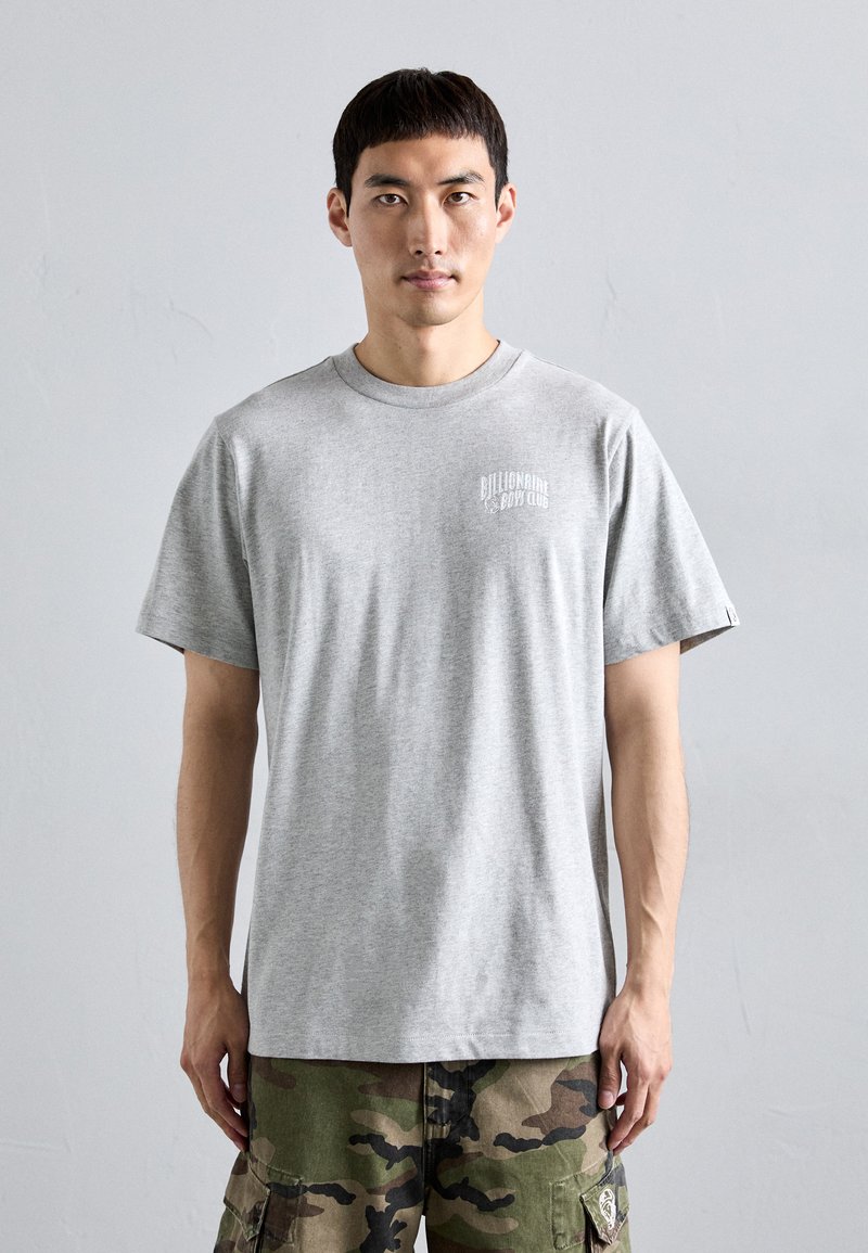 Bărbat îmbrăcat cu un tricou deshis gri de la Billionaire Boys Club și pantaloni cargo cu model de camuflaj, stând în fața unui fundal uniform de gri deschis.