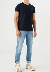 Camiseta de algodón navy con mangas enrolladas, jeans de color azul claro y zapatillas altas beige. Detalle de logo sencillo en la parte frontal de la camiseta.