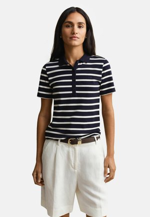Femme portant un polo à rayures bleu marine et blanches, rentré dans un short blanc taille haute avec une ceinture marron, debout devant un fond uni.