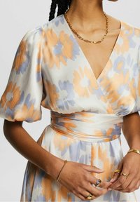 La robe portefeuille présente un motif floral orange et bleu clair sur un fond blanc, un tissu texturé, des manches bouffantes et une ceinture assortie.